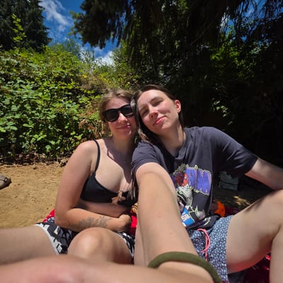 Two individuals are seated outdoors, likely on a beach or a grassy area near water, as indicated by the sandy ground and dense foliage in the background. The person on the left, wearing a black bikini top and sunglasses, has blonde hair and a tattoo on their arm. The person on the right, wearing a dark t-shirt with a graphic print, has dark hair and is looking towards the camera. They are both positioned in the lower half of the frame, with the upper portion dominated by trees and a bright blue sky with scattered clouds. The lighting suggests it is a sunny day. The overall impression is a casual, relaxed outdoor scene. It is not possible to determine the specific location within Cherry Grove, United States, from the visual information provided.