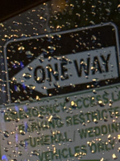 The image shows a close-up of a sign with water droplets on it, suggesting it might be raining or the sign is wet. The sign has the words "ONE WAY" in large white letters on a dark background, with an arrow pointing to the left. Below "ONE WAY", there are lines of green text that are partially obscured by the water droplets and the angle of the photo. The visible text includes "EMERGENCY ACCESS" and "PARKING RESTRICTED". Further down, parts of "FUNERAL/WEDDING VEHICLES ONLY" are visible. The background appears to be a dark, out-of-focus surface, possibly the exterior of a building or another sign. The lighting is somewhat dim, with bright reflections from the water droplets. The image does not provide enough information to determine its specific location in Stony Plain, Canada.
