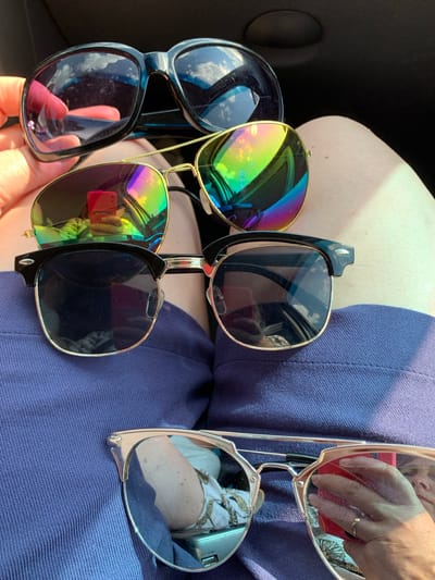 Four pairs of sunglasses are laid out on a person's lap, which is covered by dark blue fabric. From top to bottom, the first pair is black with large, rounded lenses that reflect the sky and clouds. The second pair has gold frames and lenses with a rainbow-colored reflection. The third pair has black frames with a metal brow bar and dark, reflective lenses. The fourth pair, at the bottom, has silver frames and reflective lenses. The background is dark. This image was taken indoors, likely in a car, and is not related to the city of Азерскі сельскі Савет, Belarus.