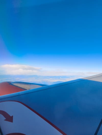 This is an aerial photograph taken from an airplane window, looking out at the sky and clouds. The airplane's wing is prominently featured in the foreground, painted in shades of blue and white with a red stripe and an arrow. The wing extends diagonally across the frame, from the bottom left to the top right. Above the wing, a layer of white clouds is visible, with the vast expanse of a clear blue sky above them. There are no people, buildings, or specific landmarks of Sebnitz, Germany, visible in this image. The scene depicts a typical view from an airplane in flight, high above the ground.