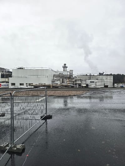 The image depicts an industrial scene in Haguenau, France, under an overcast sky. The foreground features a dark, wet asphalt surface exhibiting reflections of the overhead sky and distant structures, indicating recent precipitation. Two sections of temporary metal fencing, supported by dark rectangular bases, are positioned in the lower left and central foreground. A faint white line, possibly painted, traverses the wet ground horizontally.
In the midground, a large industrial complex extends across the frame. This complex consists of multiple white or light grey buildings, some with flat roofs and visible pipework. A prominent cylindrical stack, with a flared top, rises from the central part of the complex. To the right, another stack emits a plume of light grey smoke or steam vertically into the cloudy sky. A partial sign on a building to the left reads "LOGISTIC". Dark green trees are visible along the horizon behind the right side of the industrial buildings. No individuals are present in the scene.