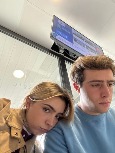 This image captures two young adults in the waiting area of Imagerie Médicale du Sud-Est Lyonnais, located at 10 Rue du Dauphiné, Saint-Priest, France. The female on the left, with blonde hair, rests her head on the right shoulder of the male, who has brown hair. She looks towards the camera with a neutral expression, wearing a tan jacket over a dark inner layer. The male looks down and to his right, away from the camera, wearing a light blue sweatshirt. Above them, a flat-screen monitor displays medical facility information, including "Doctolib," "IMAGERIE MÉDICALE DU SUD-EST LYONNAIS," and the queue number "169." The background shows a white suspended ceiling with integrated lighting and a glass partition reflecting the word "ologie" in reverse, indicating a clinical environment.