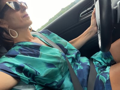 The image shows a woman driving a car. She is wearing a blue and green patterned dress and large sparkly earrings. Her hand is on the steering wheel, and she is looking to her right. The interior of the car is visible, including the dashboard and door panel. Outside the car, there is greenery, suggesting a rural or suburban setting. The time of day appears to be daytime, with bright light coming from the right side of the frame. The weather appears to be clear and sunny. The overall mood of the image is casual and relaxed.