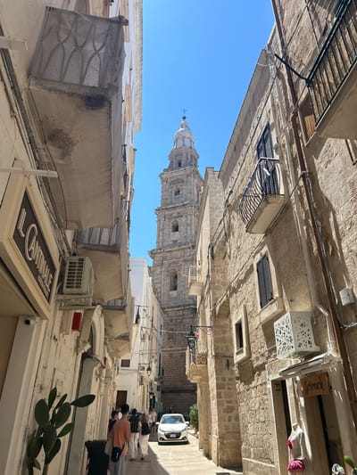 The image depicts a narrow, sun-drenched street in Monopoli, Italy, with a prominent bell tower rising in the background. The architecture is characterized by stone buildings with balconies and arched windows. On the left side, an air conditioning unit is visible on a building facade, as is a sign for "Il Cantinale." On the right side, a store window displays mannequins dressed in clothing, and a sign reads "Scrirock." Several people are walking down the street, some appearing to be tourists. A white car is parked in the distance. The sky is clear and blue.