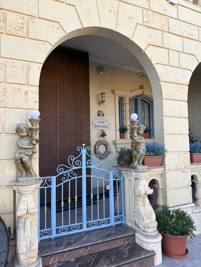 The image depicts the entrance to a building in Xagħra, Malta. The entrance is framed by a stone archway, with decorative stonework on the facade. To the left, a small statue of a cherub holding a torch with a glowing bulb stands on a pedestal. In front of the entrance is a light blue wrought-iron gate with decorative scrollwork and gold accents. A nameplate above the gate reads "CASA MARI". To the right of the entrance, another statue of a cherub holding a torch is visible, placed on a pedestal with a carved fish motif. Several potted plants, including lavender, are arranged on ledges and the ground. The building's exterior is constructed of light-colored stone, with some areas showing signs of wear. The scene is illuminated by natural daylight.