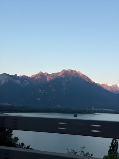 This is a vertical, medium-wide shot of a mountain range with a lake in the foreground, taken from a moving vehicle. The sky is a clear gradient of light blue to pale yellow. The mountains are dark blue in shadow, with the peaks illuminated by warm, golden light, suggesting sunrise or sunset. A thin line of trees and a few scattered buildings are visible at the base of the mountains, along the lake shore. A body of water stretches across the lower half of the image, with ripples on its surface. A dark, blurred railing runs horizontally across the middle and bottom of the frame, indicating the vantage point of the photographer is from inside a vehicle. The image appears to have been captured near Villeneuve, Switzerland, given the presence of the lake and mountains often associated with the region. There are no people or discernible actions taking place.