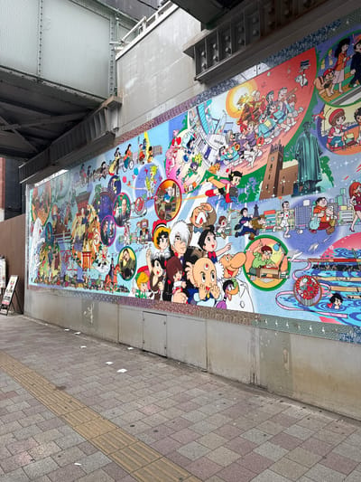 The image displays a long, vibrant mural on a concrete wall in Shinjuku, Japan. The mural depicts numerous characters from Japanese manga and anime, in a colorful and detailed style. The artwork spans the entire length of the wall, featuring scenes with many cartoon figures engaged in various activities, including playing music, running, and interacting with each other. Some characters appear to be from classic anime series, recognizable to fans of Japanese animation. The mural is located outdoors, with a sidewalk in the foreground paved with interlocking bricks. Above the mural, a portion of an overpass or elevated structure is visible, with exposed metal beams and a grey metal walkway or roof. The overall scene is a public space, likely a pedestrian area under an elevated train line or highway, decorated with artistic representations of popular Japanese culture.