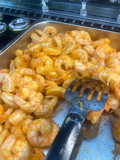 A close-up, overhead shot shows a metal tray filled with a large quantity of cooked shrimp seasoned with a reddish-orange spice mix. A portion of a metal slotted serving utensil is visible in the foreground, partially submerged in the shrimp. The shrimp appear to be cooked and ready to serve, likely in a buffet or food service setting. In the background, out of focus, are glass jars with dark contents, possibly caviar, and a metal grate with a black surface. The overall scene suggests a food display. No specific location in Khanty-Mansiysk, Russia can be determined from the image.