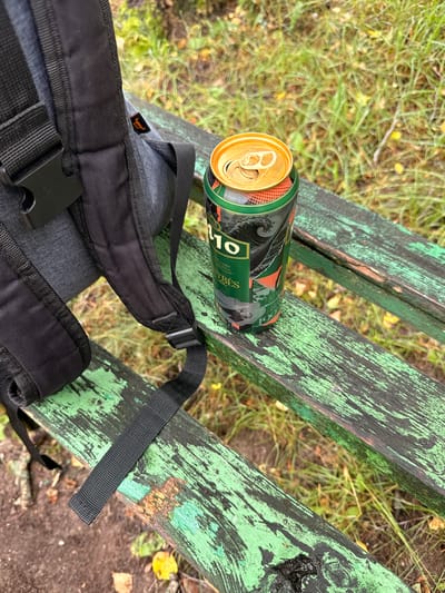 A black backpack is placed on a weathered green wooden park bench, with its straps visible. An open can of beer with a green and black design sits on the bench next to the backpack. The background consists of grass and fallen leaves, suggesting an outdoor setting. The scene is located in Rudamina, Lithuania.