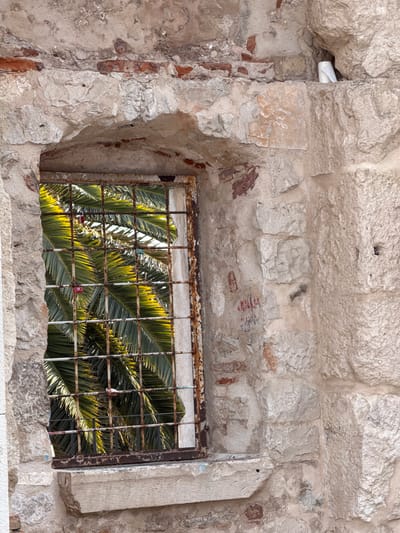 The image shows a rough, weathered stone wall with a partially broken opening that serves as a window. The window is covered by a rusty metal grate, behind which palm fronds are visible, backlit by a bright light, suggesting it is daytime. The stone wall is made of irregular blocks of varying sizes, with visible signs of age and decay, including patches of red brick and crumbling mortar. The opening in the wall is jagged and uneven, highlighting the dilapidated state of the structure. Some faint red markings, possibly graffiti, are present on the stone above the window. The overall scene evokes a sense of abandonment or neglect in a warm climate, potentially a historic site in Split, Croatia, given the palm trees.