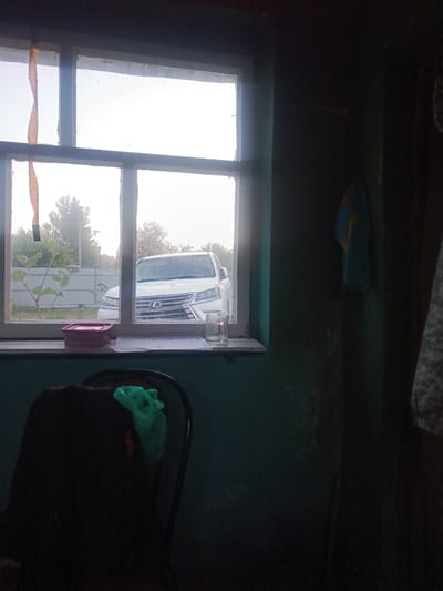 The image depicts an interior view looking out a window. The window frame is weathered and divided into multiple panes. Outside, a white Lexus SUV is parked behind a fence. Green foliage and trees are visible in the background under an overcast sky. On the windowsill, a small pink container, a glass, and some papers are present. Inside, in the foreground, a dark chair with a crumpled green cloth draped over its back is partially visible. The interior walls appear to be painted a dark green color, and a blue and yellow towel hangs on the wall to the right. The scene is dimly lit, suggesting it might be daytime but with low light conditions or the interior is not well-lit. The overall impression is one of a humble, possibly rural or semi-urban setting in the city of Poshekhonye, Russia.