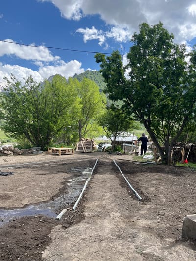The image depicts an outdoor scene with a focus on a dirt path lined with young trees and under construction. The foreground shows a muddy area with water flowing through it, bordered by newly laid concrete curbs. Two parallel lines of white pipes are visible, suggesting an irrigation or drainage system installation. Further back, a person is standing near a structure, facing away from the camera. Several wooden pallets are stacked on the left side of the path, and a pile of wood is visible on the right. In the background, lush green trees and mountains under a partly cloudy blue sky frame the scene. The overall environment suggests a rural or mountainous setting, consistent with the description of Sioni, Georgia.