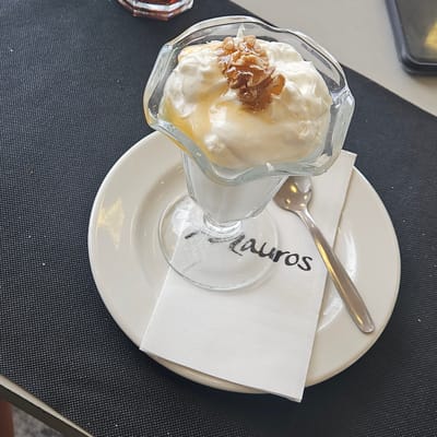 This is a close-up, high-angle shot of a dessert served in a glass bowl on a white plate, placed on a dark textured placemat. The dessert appears to be thick, white yogurt or cream, drizzled with honey and topped with a cluster of what looks like caramelized nuts or candied fruit. A silver spoon rests on the white napkin and plate to the right of the dessert bowl. The glass bowl has a logo or text on its base which reads "Lauros". The overall scene suggests a casual dining setting, likely a cafe or restaurant. The location is identified as La Font de la Figuera, Spain. There are no people visible in the image.