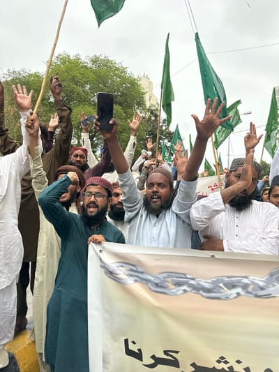 In Karachi, Pakistan, a group of men are holding green flags and a banner, with some raising their hands in the air. One man is holding a smartphone. The men are dressed in traditional Islamic clothing. The scene appears to be a protest or a rally, with a backdrop of trees and buildings. The weather is overcast.