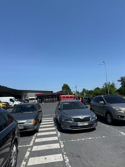 The image captures a bright, sunny day at a parking lot in the city of Sarny, Ukraine. In the foreground, several cars are parked on a gray paved surface marked with white pedestrian crossing lines. A dark gray sedan is visible on the left, partially cropped, with its rear wheel and fender in view. Next to it, a light gray sedan is parked, facing forward, with its license plate readable as "1 BA". Further to the right, a silver Skoda Superb is parked, also facing forward, with its license plate showing "UA AT 9651 IA". Behind it, a silver Hyundai SUV is parked. In the middle ground, a small storefront with a red awning and signage is visible, displaying "ФРУКТИ" (Fruits) and "Овочі" (Vegetables). Several people are seen near the storefront, some appearing to be shoppers. A red mini-bus is parked to the left of the storefront. In the background, industrial-style buildings with gray roofs and concrete block walls are present. Trees and power lines are visible in the distance, under a clear blue sky. The overall scene depicts a commercial area with a parking lot and a small market.