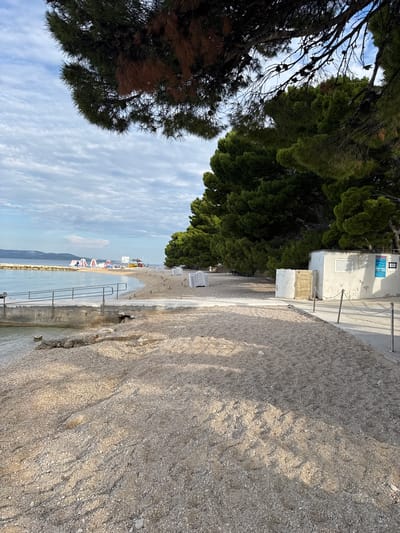 A pebble beach in Makarska, Croatia, is partially shaded by tall pine trees. A concrete walkway runs along the edge of the beach, with a railing for support. The water is calm and shallow near the shore. Scattered on the beach are rows of white lounge chairs, suggesting a tourist area. In the background, there are some inflatable water toys and a small white building. The sky is partly cloudy.