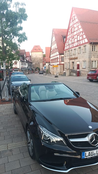A black Mercedes-Benz convertible is parked on a cobblestone street in front of traditional German timber-framed buildings. The car's interior, with light-colored leather seats, is visible. To the left of the convertible, other cars are parked, including a dark SUV. In the background, a historic town gate stands with more half-timbered houses and a tower. A red vehicle is parked further down the street. A yellow sign with the word "Umlaufung" is also visible. The scene appears to be set in Altdorf bei Nürnberg, Germany, during daylight hours, possibly late afternoon given the soft light. The atmosphere is calm and picturesque. The license plate of the Mercedes-Benz reads "LAU L....". A sign on the building reads "Raiffeisenbank". Another sign on a building on the left shows "Moeller".