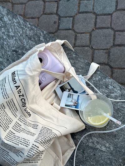 An overhead shot shows a beige tote bag resting on a weathered gray stone surface, possibly a ledge. Inside the bag, among the folds of fabric, is a lavender instant camera, two small instant photos, and the corner of what appears to be a travel guide ("Milan A to Z City Guide"). A clear plastic cup filled with a clear liquid, possibly lemonade, with a straw and a lemon slice sits on top of the bag, along with a white charger cable. The setting appears to be outdoors, with cobblestones visible to the side of the ledge. Gittana, Italy is not mentioned in the text of the bag.