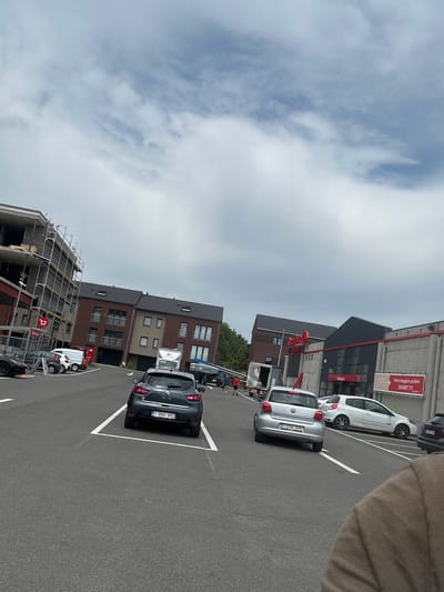 The image captures a street scene in Herstal, Belgium, on an overcast day. The perspective is from a person standing on a paved area, likely a parking lot, looking down a row of parked cars and buildings. Several cars are visible, including a silver Volkswagen with license plate "1 EXG-895" and a dark gray Renault Clio with license plate "1-BWD-893." In the background, modern brick buildings with dark roofs line the street, interspersed with construction sites featuring scaffolding. One building displays a red sign that reads "Votre magasin préféré OUVERT 7/7," indicating it is a shop open seven days a week. The sky is filled with diffused light from the clouds, suggesting a cloudy or partly cloudy day. The overall impression is a typical urban environment with ongoing construction and commercial activity.