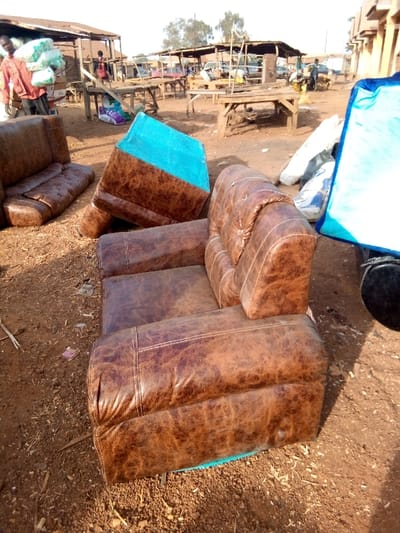 An outdoor scene displays a dissembled brown leather couch with visible blue fabric underneath, set on reddish-brown dirt. A two-seater couch sits in the background. People and market stalls are seen at a distance. A blue covering lies to the right, partially obscuring a dark object. The area appears to be a commercial or market setting.