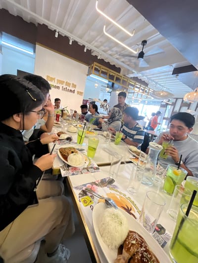 The image depicts a group of people enjoying a meal at a restaurant. The restaurant appears to be modern with white walls, striped wall panels, and contemporary lighting fixtures. A ceiling fan is visible. The tables are set with white placemats featuring a tropical design and text that reads "Wake up to Delicious Cinnamon Island way!". White plates with rice and what appears to be glazed chicken are prominently displayed. Several glasses of a green beverage, some with ice and lemon slices, are on the table, indicating drinks are being served.
There are approximately eight individuals visible, some of whom are actively eating or drinking. A young woman in the foreground is looking at her phone while holding a spoon and fork. Other diners are engaged in conversation and eating their meals. In the background, two individuals are standing, and other patrons are visible at different tables.
The overall impression is of a lively and casual dining experience. The restaurant's decor and the food served suggest a potentially tourist-oriented establishment, possibly serving Asian cuisine. Based on the style and atmosphere, this restaurant could be located in the city of Koronadal, Philippines, a city known for its culinary scene and growing tourism.