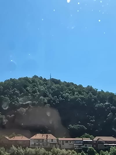 The image shows a view of a hillside covered in lush green trees under a clear blue sky. At the very top of the hill, a tall, slender structure, likely a telecommunications tower or antenna, is visible. Below the treeline, a row of houses with reddish-brown roofs lines the lower part of the hillside. The houses appear to be multi-story residential buildings with varying architectural styles. Some of the houses have visible windows and balconies. The overall scene suggests a suburban or semi-rural setting. The presence of the tower on the hill could indicate a strategic location for broadcasting or communication in the city of Zvornik, Bosnia and Herzegovina. The bottom of the image shows some greenery, possibly gardens or trees in front of the houses. There are some white specks in the sky, which could be dust, pollen, or artifacts from the lens or sensor.