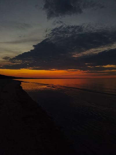 This media file depicts a tranquil scene of a beach at sunset in Apšuciems, Latvia. The image focuses on the meeting of the sand and the sea under a dramatic sky.

The primary subjects are the natural elements: a dark, sandy beach occupies the left foreground, stretching into the distance along a wooded coastline. The sea is a vast expanse of dark, calm water, reflecting the vibrant colors of the sunset. Small, gentle waves lap at the shore, leaving a shimmering wet sheen on the sand. The sky is a canvas of deep blues and grays at the top, transitioning to a brilliant band of orange and yellow near the horizon, indicating the sun is setting. Dark, textured clouds are scattered across the upper portion of the sky.

There are no people or discernible objects in the frame, emphasizing the serene and natural beauty of the location. The activity is the passive observation of a sunset, a moment of quietude and natural spectacle.

Notable details include the time of day, clearly sunset, characterized by the intense hues in the sky and the low light. The weather appears calm, with still water and soft waves. The presence of a sandy beach and the Baltic Sea is implied by the location context. The overall emotion evoked is one of peace, serenity, and the awe-inspiring beauty of nature. There is no visible text in the media file.