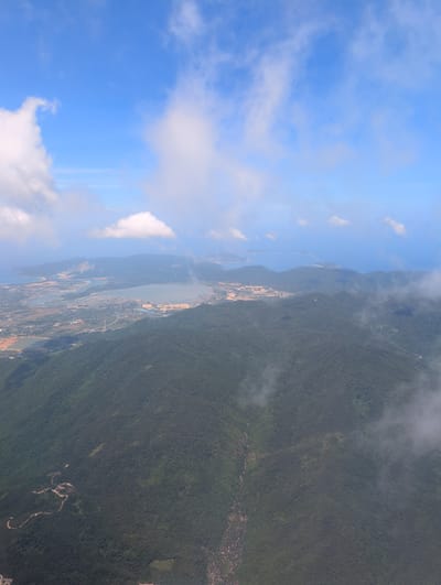 This is an aerial photograph of a mountainous, coastal landscape with a bright blue sky dotted with white clouds. The foreground is dominated by a lush, dark green, densely forested mountain that slopes downward towards a body of water. A winding, light-colored dirt road snakes its way across the face of this mountain, disappearing into the vegetation.

In the middle ground, a large, calm lake or bay is visible, with lighter green and tan land surrounding it. Various structures and signs of development, including what appear to be industrial sites or construction areas, are scattered around the lake's perimeter and in the surrounding plains. There are also some smaller bodies of water and canals. Distant, lower-lying mountains, also covered in vegetation, form the backdrop.

The horizon shows the ocean meeting the sky, with a few scattered islands or peninsulas in the far distance. The lighting suggests it is daytime, likely late morning or early afternoon, with bright sunlight casting some shadows. The overall scene depicts a developing coastal region where natural mountain and water features are integrated with human infrastructure. This landscape is consistent with the geography of Sanya City, China, known for its tropical climate, mountains, and coastal development.