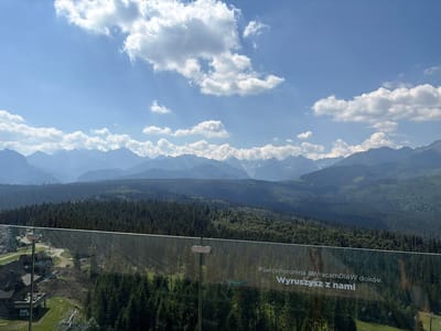 This image captures a breathtaking panoramic view of the Tatra Mountains in Małe Ciche, Poland, under a bright, partly cloudy sky. The scene is observed from a vantage point behind a clear glass barrier, offering an unobstructed vista of rolling forested mountains and distant, snow-capped peaks.
In the foreground, the glass barrier is slightly reflective, showing a faint image of the dense pine forest below and the sky above. The text "#SercePoronina #WracamDlaW doków Wyruszyusz z nami" is printed in white on the glass, suggesting a promotional message related to the location or an event.
Below the barrier, a charming village with traditional wooden houses and chalets is nestled on a green hillside. A ski lift infrastructure is also visible, hinting at the area's popularity as a tourist destination, likely for both winter sports and summer mountain activities.
The landscape is characterized by lush green forests that blanket the slopes of the mountains, transitioning to rugged, rocky peaks in the distance. The time of day appears to be midday or early afternoon, given the bright sunlight and the sharp shadows cast. The weather is pleasant, with scattered white clouds drifting across a vibrant blue sky. The overall mood is serene and majestic, inviting the viewer to experience the beauty of the Polish mountains.