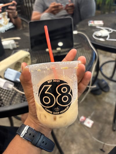 A close-up, eye-level shot captures a hand holding a clear plastic cup filled with an iced, light-colored beverage, likely coffee or milk tea. Condensation is visible on the outside of the cup, suggesting a cold drink in a warm environment. A red straw is inserted into the drink. The cup features a black circular label with the prominent white numbers "88" and smaller text reading "COFFEE SHOP PREMIUM" curved beneath, and "EST 2021" curved along the left edge.
The hand is wearing a black wristwatch with blue stitching and a silver buckle. The background is softly blurred, revealing a casual setting. A black mesh table surface is visible, upon which lies a black tablet or laptop and several white charging cables. Further back, partially obscured, another individual in a grey shirt appears to be seated at the table, possibly engaged with their own electronic device. A beige power strip or outlet is also visible on the table. The ground, visible beneath the table, appears to be a textured concrete surface, with a pair of dark shoes or slippers blurry in the distance. The overall scene suggests a relaxed atmosphere, possibly at a coffee shop or cafe in Keudeublang, Indonesia, where individuals are enjoying refreshments and utilizing electronic devices. The lighting indicates it is daytime.