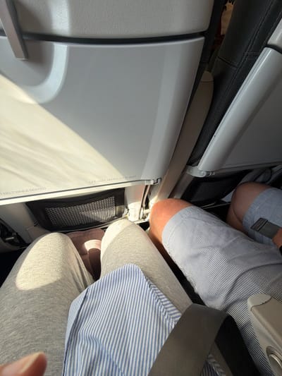 The image captures an interior view of an airplane cabin, looking forward from a passenger's seat. Two individuals are partially visible, occupying seats in front of and next to the viewer. In the foreground on the right, a person is seated, wearing a blue and white striped shirt and light grey sweatpants, with a dark seatbelt across their lap. Their lower torso and legs are visible, with a small portion of a hand or finger in the bottom right corner. Directly ahead, the knees and lower thighs of another person, clad in a blue and white striped shirt and light shorts, are seen between the seat in front and the person on the right.
The seatback directly in front of the viewer features a closed white tray table, which displays inverted text: "FASTEN SEAT BELT WHILE SEATED" and partially "LIFE VEST UNDER YOUR SEAT." A black mesh storage pocket is affixed to the seatback, visible between the tray table and the second person's legs. Bright, direct light, possibly sunlight, illuminates the cabin, creating strong highlights and shadows on the surfaces and clothing. The setting is an airplane interior, consistent with the provided location context of Spata-Loutsa Municipal Unit, Greece, indicating an airport or in-flight scene. The activity shown is simply people seated in an airplane, likely during a flight or boarding.