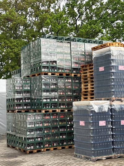 The image displays an outdoor logistics area in Valmiermuiža, Latvia, featuring extensive stacks of bottled beverage crates and empty pallets situated on paved ground beneath an overcast sky. On the left, multiple towering formations of green plastic beverage crates, secured with clear plastic shrink wrap, rise approximately twelve to fourteen crates high on wooden Euro-pallets. Each crate holds numerous small, dark glass bottles distinguished by various colored labels. To the right, several stacks of empty wooden Euro-pallets stand alongside large, shrink-wrapped stacks of dark plastic crates, some displaying small pink labels and topped with white protective plastic sheeting. Lush green deciduous trees dominate the background, while the foreground consists of textured paving. No personnel are visible; the scene indicates a temporary storage or dispatch point for a beverage operation.