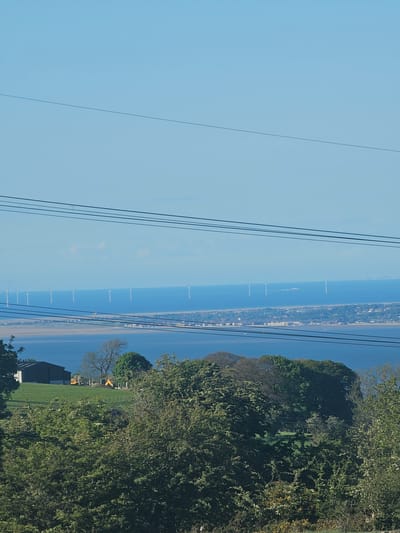 The image depicts a landscape in Rhes-y-cae, United Kingdom. The scene is viewed from an elevated position overlooking the water, a coastline with a town or village, and the surrounding countryside. High-voltage power lines are visible across the upper left of the frame. On the right, there is dense vegetation including trees and shrubs. A field or pasture can be seen along with a building and machinery such as a tractor.
