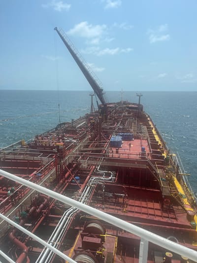 This image depicts a large marine tanker vessel at sea off the coast of Orimedu, Nigeria, viewed from a higher deck of an adjacent vessel. The tanker features a reddish-brown deck extensively covered with red and silver-colored cargo piping, valves, and platforms. A dark-colored jib crane is positioned on the port side, angled upwards and forward, with a cable and hook extending downwards. On the midship deck, two human figures are present near several blue tarpaulin-wrapped bundles and a dark blue cylindrical object. A "SAFETY FIRST" sign is visible on a deck structure. A floating boom or hose extends from the tanker's port side into the choppy blue water. The foreground shows white railings and a dark deck of the observer's vessel, including two large winches or capststans with coiled ropes. The sky is clear with scattered clouds over the open ocean.
