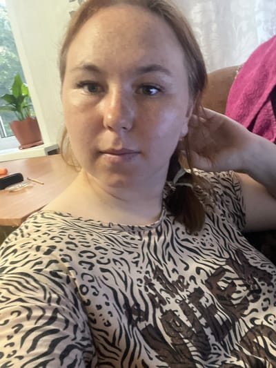 A woman is taking a selfie in what appears to be a residential indoor setting. She has fair skin with freckles, light brown hair pulled back with a ponytail, and is wearing a patterned top with a leopard and zebra print. She is sitting down, with her right hand touching her hair. In the background, there is a window with greenery visible outside, a table with a few objects on it, and a pink blanket draped over a piece of furniture. The lighting suggests it is daytime, possibly afternoon. The image does not provide any specific details about the city of Аряж, Russia.