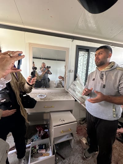 The scene is an interior room exhibiting significant damage, located in Dura, Palestinian Territory. In the mid-ground right, a man with a dark beard and short hair, wearing a grey hooded sweatshirt with "XT-FIVE" branding and black track pants, gestures with open palms as he speaks; a small microphone is clipped to his sweatshirt. To the foreground left, another individual, partially visible, holds a blue smartphone oriented vertically, appearing to record or photograph the scene. This person also wears a light brown jacket and has a professional Canon camera with a large lens slung over their shoulder; a gold wedding band is visible on their right ring finger.
Centrally positioned is a light-colored vanity or dresser with several drawers, its surface and drawers covered in dust and debris. One lower drawer is open, revealing various items including what appear to be cosmetics and a small stack of currency notes. Above the vanity, a rectangular mirror with a light-colored frame is cracked and reflects multiple individuals, including a person holding a professional camera with a flash, and other indistinct figures.
The floor throughout the room is strewn with widespread debris, including shattered glass, fragments of what appears to be a window frame or blinds, dark granular material, and distinct reddish stains. A pair of shoes, one light-colored and one dark, lie among the debris near the gesturing man. In the background right, a doorway or window opening shows severely damaged and shattered panes, further indicating structural damage to the environment. The walls and ceiling are composed of light-colored panels, with dark structural beams visible along the ceiling line.