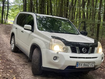 A white 2007-2013 Nissan X-Trail is parked on a dirt road in a forest, likely in the village of Kolubaivtsi, Ukraine. The car is facing the viewer at a slight angle, with its front passenger-side door ajar. The vehicle has a dark-colored hood protector. The license plate reads "BX 8662 IC" with the Ukrainian flag symbol. The surrounding environment is dense with tall trees and foliage, with sunlight filtering through the leaves, creating a dappled effect. The ground is covered with dirt, small stones, and fallen leaves. There are no people visible in the image.