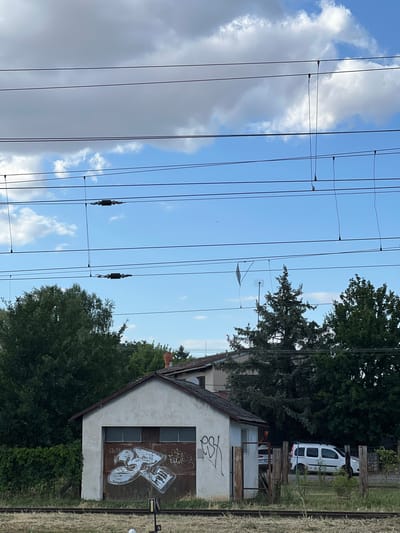 The image captures a scene in Újezd u Brna, Czechia, focusing on a small, weathered, light-colored building with a rusty corrugated metal garage door adorned with white graffiti. The building is flanked by lush green foliage on the left and a chain-link fence with wooden posts on the right. Behind the building, several trees are visible, including a tall pine and other deciduous trees, with glimpses of additional buildings and a chimney peeking through. A white van is parked to the right of the building, partially visible behind the fence. Above the scene, multiple overhead power lines stretch horizontally across the frame, suspended against a bright blue sky with scattered white clouds. The ground in the foreground consists of gravel and railway tracks.
