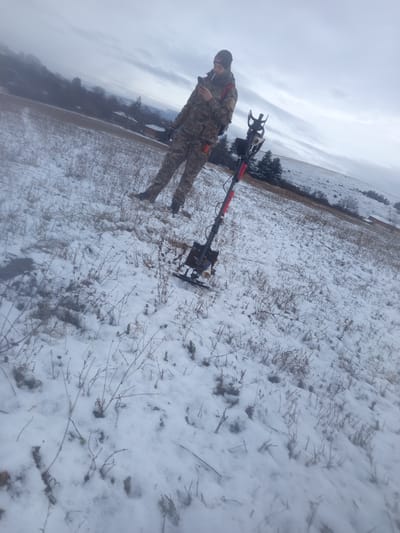 The scene depicts an individual standing in a snow-covered field located near Drugan, Bulgaria, under an overcast sky. A single adult male, dressed in camouflage clothing, a black beanie with a "Minelab" logo, dark gloves, and dark boots, occupies the central left portion of the frame. He is holding a smartphone in both hands, looking down at the screen, and has a lit cigarette in his mouth. A black and red metal detector stands upright in the snow to his right, with its search coil resting on the ground. The field exhibits a thin, patchy layer of snow, revealing dry, brown vegetation and some darker earth underneath. In the background, gently sloping terrain with sparse trees and a few distant structures are visible beneath the grey, cloudy sky.