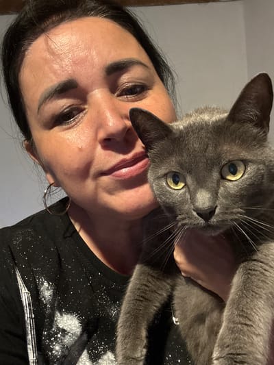 In this media file, a woman is holding a grey cat close to her face, creating an intimate selfie. The woman has dark hair and is wearing makeup, including eyeshadow and lipstick. She is also wearing a black t-shirt with a speckled white design on it. The cat has bright yellow eyes and is looking directly at the camera. The background is a plain, light-colored wall, suggesting an indoor setting. The lighting appears to be artificial, casting shadows and highlighting the subjects. The image captures a moment of affection between the woman and her pet, with both subjects displaying a relaxed and affectionate demeanor. No specific time of day or weather conditions are discernible from the image. The location is identified as Starokhalilovo, Russia.