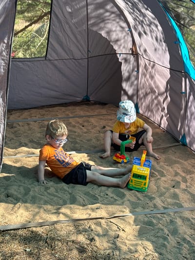 Two young children are playing in a sandpit within a tent. The child on the left, wearing an orange t-shirt and black shorts, is sitting with their legs outstretched. They have light-colored hair and are wearing glasses. The child on the right, wearing a yellow t-shirt, black shorts, and a patterned bucket hat, is kneeling and playing with two toy trucks, one green and yellow, the other yellow and blue. The sandpit appears to be an outdoor location, possibly in Ozerki (Krasnodolinskaya), Russia, with trees and foliage visible through the tent opening. The tent itself is made of grey fabric with blue accents. Sunlight casts dappled shadows on the sand and tent.