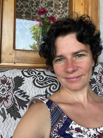 A medium shot portrait of a woman with short, dark, curly hair and blue eyes is taken indoors. She is looking directly at the camera and smiling. Her skin is fair with some redness on her cheeks and nose, suggesting sun exposure. She is wearing a patterned top with floral and geometric designs in shades of blue, purple, white, and orange. She is seated on a couch with a similarly patterned, ornate fabric. Behind her, a wooden framed window with white lace curtains is visible. Through the window, trees and sky are reflected. The scene appears to be in a residential setting within the Ghvris Khevi community in Georgia.