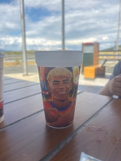 A McDonald's cup with an image of Kylian Mbappé is placed on a wooden table outdoors, with a blurred background of a McDonald's restaurant and a bright sky. The cup is white with a colorful illustration of the soccer player smiling and wearing braces. The table has visible grain and a gray inlay. To the left of the cup is another white cup with some labeling. In the background, there's a blurry view of a fast-food restaurant building with orange and red elements, and some seating areas. The sky is bright with scattered clouds, suggesting an outdoor setting. The image is likely taken in Drazhevo, Bulgaria, a location known for its McDonald's restaurants.