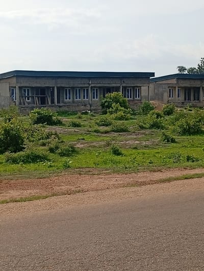 Two unfinished, single-story buildings are situated in a clearing with overgrown vegetation. The buildings appear to be under construction, with exposed brickwork and unfinished window frames. The larger building to the left has multiple windows and an open entrance. The smaller building to the right also has windows. The surrounding environment is a mix of dry earth and green grass with scattered bushes. A paved road runs along the bottom of the image. The sky is overcast. The scene is located in Shuwa, Nigeria.