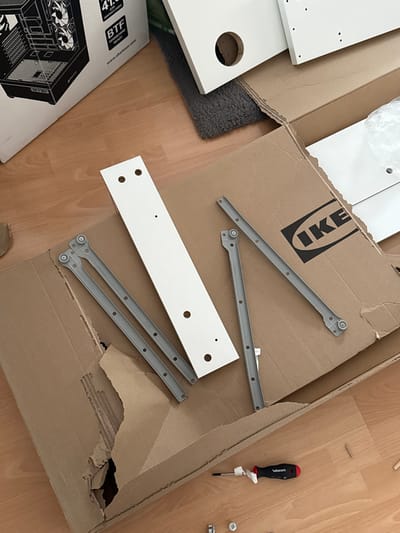 The scene is an indoor location in Vellmar, Germany, depicting a collection of unassembled furniture components and computer hardware packaging laid out on a light brown wooden floor. In the center, a large, open, brown cardboard box, featuring the "IKEA" logo on its inner flap, is visible. This box appears partially torn on its left side. Within and around the box are several white, flat-pack furniture panels of various sizes, drilled with multiple holes for assembly. Prominently featured on the cardboard box is a long, narrow white panel with multiple circular and smaller drilled holes. Adjacent to this, multiple grey metal drawer slides, each with small wheels at one end and screw holes along their length, are distributed.
In the upper left portion of the image, a white and black cardboard box for a "Sharkoon TG4M RGB" computer case is present, displaying an illustrative rendering of the product and text including "41.9 cm" and "BTF." A dark grey, shaggy rug is partially visible beneath some of the white panels and the IKEA box in the upper central area. On the wooden floor in the lower right, a screwdriver with a black and red handle, marked "WIESMANN" and adorned with a white plastic tie, lies next to several small wooden dowels and various metal fasteners, including screws and cam locks. No individuals are present in the scene.