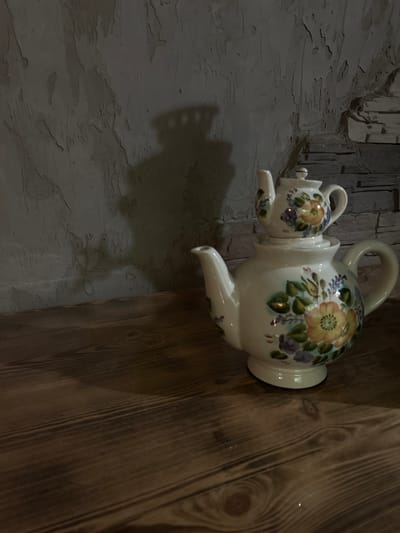 A decorative, white ceramic teapot with a smaller teapot stacked on top sits on a dark wooden surface. Both teapots are adorned with painted flowers in shades of yellow, orange, purple, and green. The teapots are positioned in front of a textured, grey wall with a brick pattern visible on the right side. A shadow, cast by the teapots and possibly an unseen light source, is projected onto the wall. The scene is dimly lit, with the focus on the teapots. The location is the city of Slov'yanoserbs'k, Ukraine.