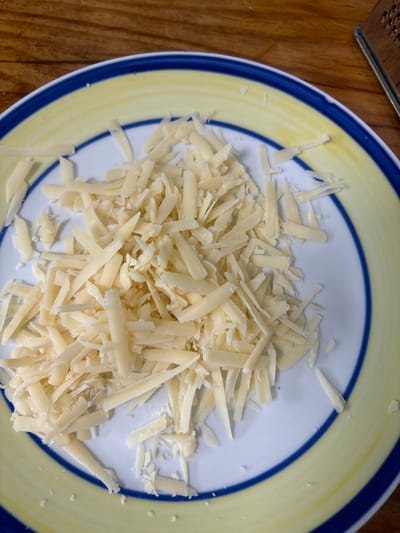 A white ceramic plate, featuring concentric yellow and blue bands along its rim, is centered with a dense pile of light yellow, irregularly shredded cheese. The cheese shreds vary in length and thickness, with some smaller fragments dispersed around the main mass. A stainless steel box grater, specifically its side with small, raised grating surfaces, is partially visible in the upper right corner of the frame. The background consists of a brown wooden surface, likely a countertop or cutting board, visible in the upper portion of the image. No individuals are present. The scene is located in the Menashe Regional Council, Israel.