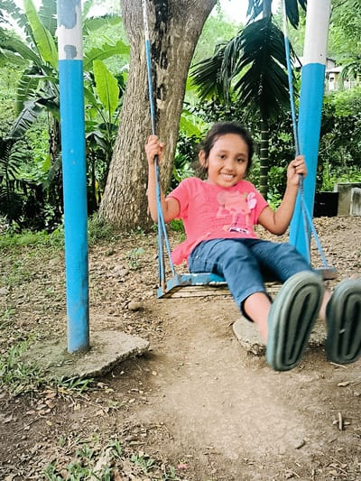 A young girl, likely Asian, is captured mid-swing on a simple, rustic swing set in an outdoor, verdant setting in Lambarotunong, Indonesia. She is the central subject, radiating joy with a wide, bright smile. The girl wears a pink t-shirt featuring a cartoon character graphic, blue jeans, and dark green sandals; her feet show motion blur, indicating she is actively swinging forward.
The swing set consists of two blue and white painted metal posts supporting a blue wooden seat. Behind the swing, the background is dominated by lush tropical foliage, including prominent banana leaves and palm fronds, as well as the thick, textured trunk of a large tree. The ground is a mix of dry earth, fallen leaves, small rocks, and sparse green grass, with a visible concrete base anchoring one of the swing posts. The natural, soft light suggests it is daytime, either morning or late afternoon. The scene evokes a feeling of simple happiness and play in a natural, tropical environment. No distinct text is clearly legible other than the character on her shirt.