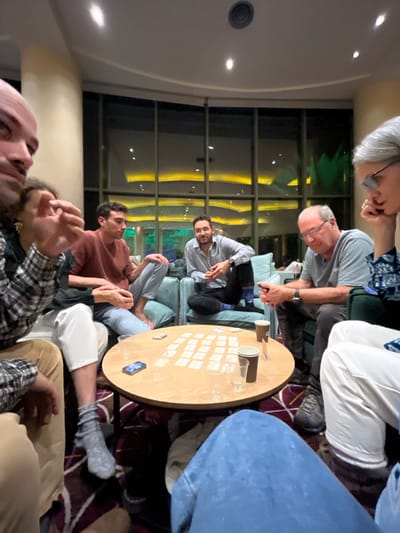 This image, rotated 90 degrees clockwise, captures an indoor social gathering of several adults at night, likely within a lounge or common area in Ein Bokek, Israel.
The scene features a group of approximately seven to eight individuals of varying ages, dressed in casual attire, seated around a light wooden, round table. On the table, numerous small, white rectangular cards are neatly arranged in rows, suggesting a card or board game is actively being played. Also on the table are two paper cups and a smartphone or similar device with a blue screen.
Among the people, a man with a beard and patterned shirt sits on a light blue couch, looking directly towards the viewer. An older man with glasses and short grey hair is seated nearby, focused on the table. Another younger man in a reddish-brown sweater also looks towards the table, while other partial figures with varying hair colors (white/grey, dark) are visible in the foreground and background, engaged in the activity or conversation.
The interior is softly lit by recessed ceiling lights. Dominating one wall are large, dark-framed windows that reveal the dark exterior of night. Through these windows, distant outdoor lights with a golden glow are visible, along with the faint silhouettes of palm trees, hinting at a warm, resort-like environment. The overall atmosphere appears relaxed and convivial, indicating a pleasant social event. No discernible text is visible on any objects or cards.