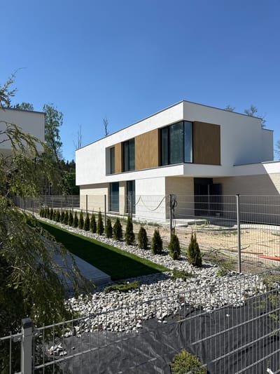 The image depicts a modern, multi-story building in Wiązowna, Poland, under a clear blue sky. The building features white walls with wooden accents around the windows. A line of small, evergreen trees borders the building, adjacent to a landscaping feature composed of grey rocks and a black geotextile fabric, secured by a wire fence. A green garden hose is visible near the building's facade. The setting suggests a newly developed or landscaped residential area.